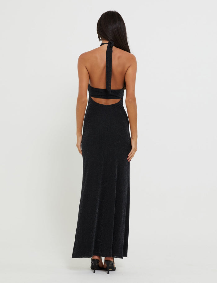 A woman with long dark hair stands facing away, showcasing the sleek, sleeveless TIANAH MAXI DRESS - BLACK from TIGERMIST. The halter neck gown reveals her back and features a cutout detail just below the shoulder blades. She pairs the floor-length dress with black heels and stands against a neutral background.