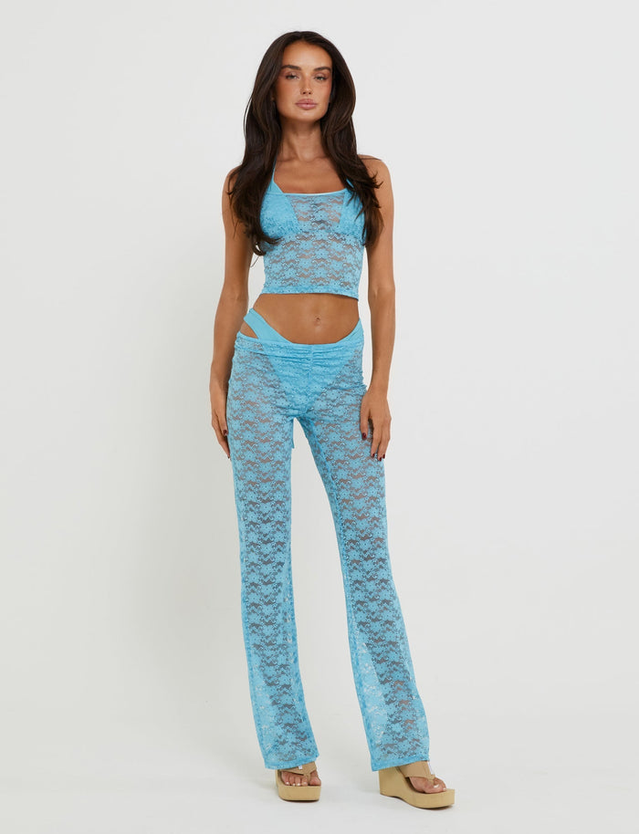 A woman stands against a plain white background wearing the TIGERMIST Tobias Pant in ice blue, featuring a bright blue lace design. The ensemble consists of a sheer, light blue sleeveless crop top paired with high-waisted, wide-legged pants. She has long, dark hair and is wearing beige platform sandals.