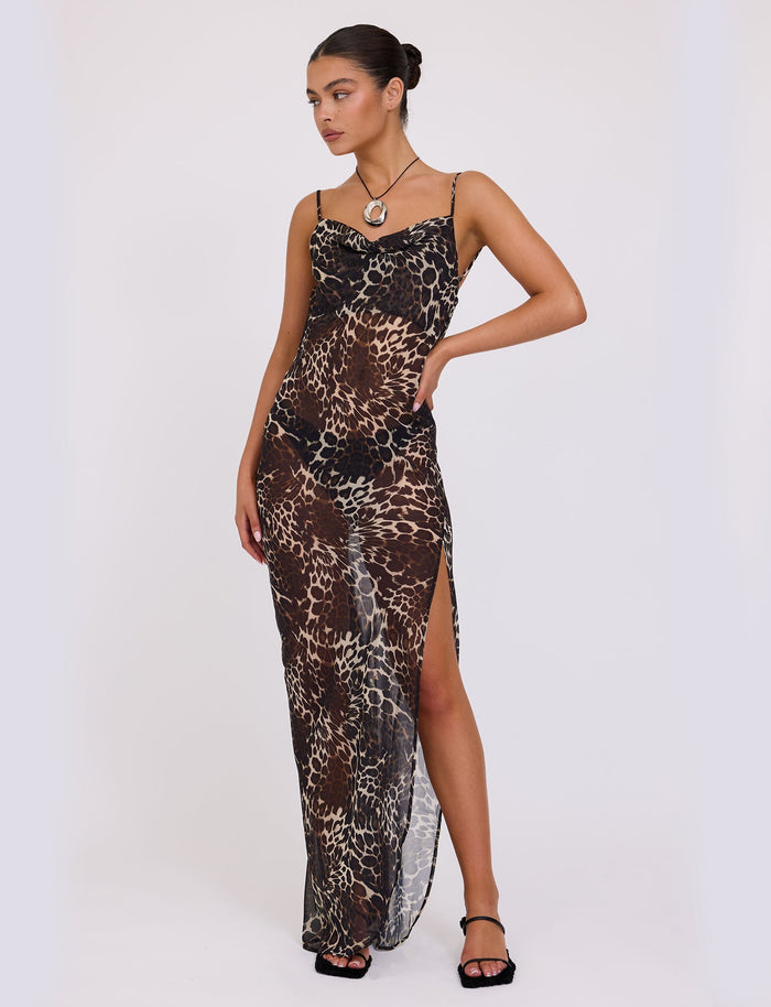 A woman stands against a white background wearing the sleeveless TYNE DRESS - MULTI: LEOPARD maxi dress from TIGERMIST, featuring a thigh-high slit. Her hair is styled in a bun, she accessorizes with a pendant necklace, and she wears black strappy heels. She poses with one hand on her hip.