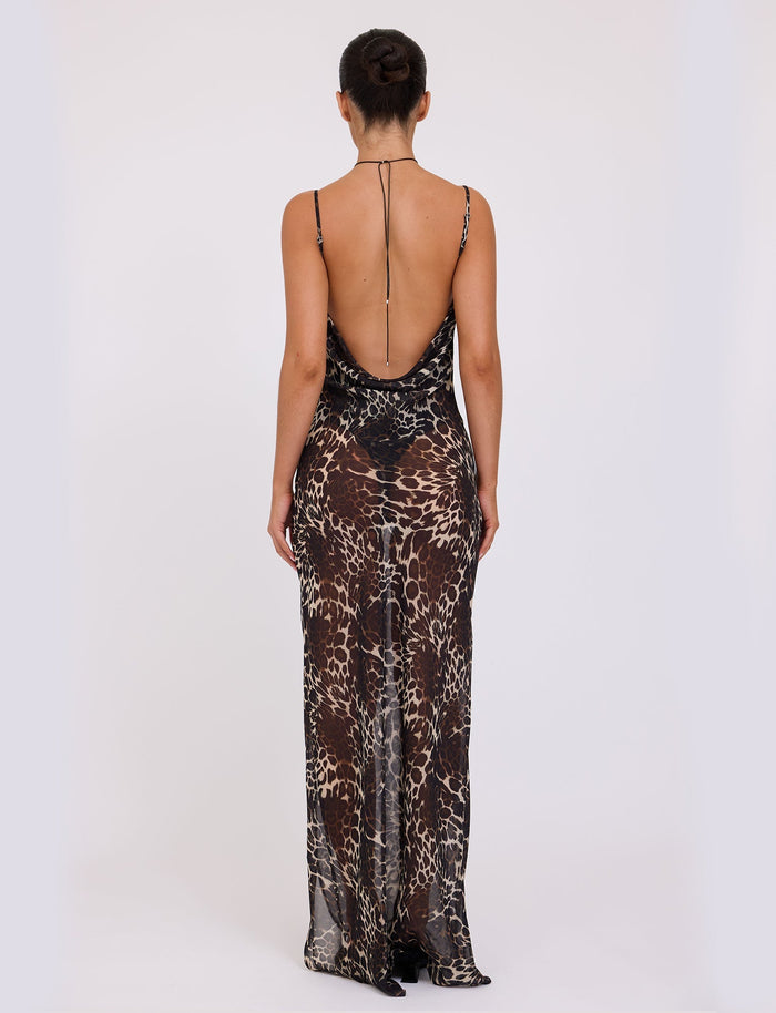 A person stands with their back to the camera, wearing the TYNE DRESS - MULTI: LEOPARD from TIGERMIST. The open back of the long, sleeveless, animal print dress reaches down to their lower back. The dress is fitted and flows gracefully down to the floor, while their hair is styled in a neat bun.