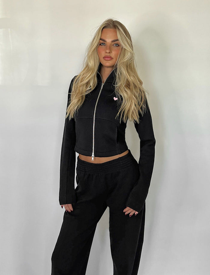 A person with long blonde hair is standing against a plain white background. They are wearing the VERDA JACKET - BLACK from TIGERMIST along with matching black pants. Their hands rest at their sides, and they have a neutral expression on their face.