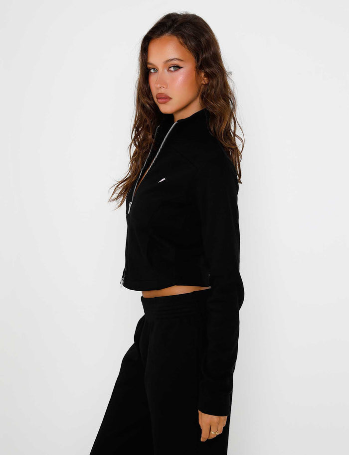 A woman with long, wavy brown hair is standing against a plain white background. Clad in the sleek black VERDA JACKET by TIGERMIST and matching pants, she gazes directly into the camera with a neutral expression. Her right hand rests at her side.