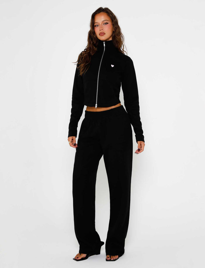 A woman stands against a plain white background, wearing the VERDA PANT - BLACK from TIGERMIST. She showcases a stylish black Tiger Mist outfit that includes a cropped zip-up jacket featuring a small heart detail paired with high-waisted wide-leg pants. With her long, wavy brown hair cascading down, she strikes a pose with one hand on her hip.