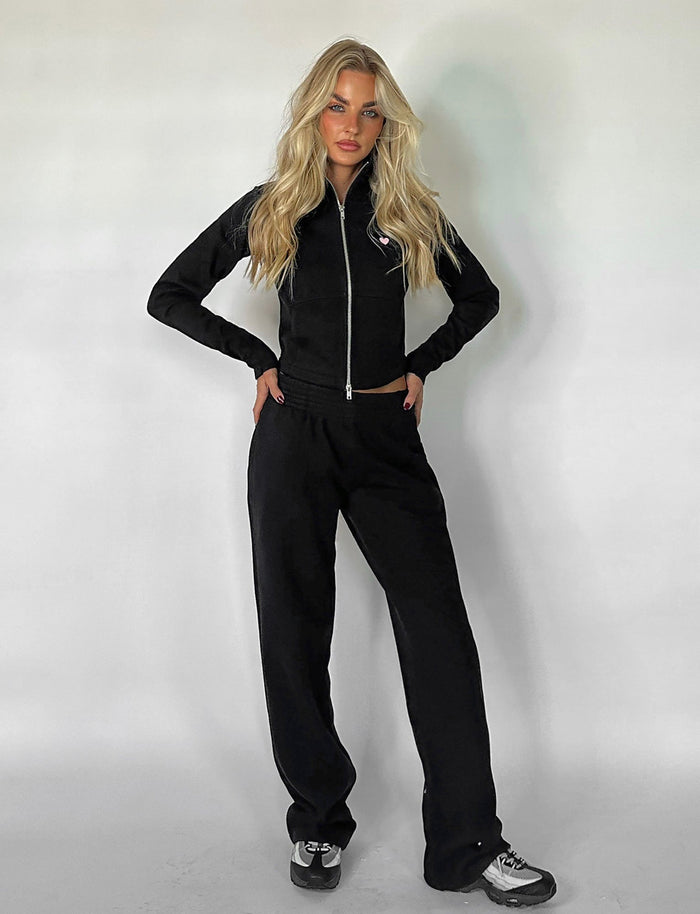 A person with long blonde hair stands against a white background, wearing a sleek Tiger Mist black zip-up jacket and matching VERDA PANT - BLACK from TIGERMIST. They are posing with their hands on their hips and sporting stylish black and white sneakers.