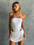 A woman poses against a textured gray wall wearing the TIGERMIST VERITY SKORT in white, styled with a strapless top, her hands holding the skort as she looks slightly to the side.