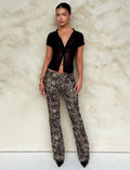 A young woman stands against a neutral background wearing a black short-sleeved, semi-sheer button-up top paired with the TIGERMIST VESPER PANT - MULTI : LEOPARD. She accessorizes with gold bracelets and earrings, and her hair is pulled back into a sleek bun.