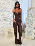 A woman with long dark hair wearing a strapless deep neckline top and the WILDER PANT in RED BROWN from TIGERMIST stands in a stylish room. Behind her, a wavy full-length mirror, a white abstract wall, and a small sculptural table are visible. She gazes confidently at the camera.
