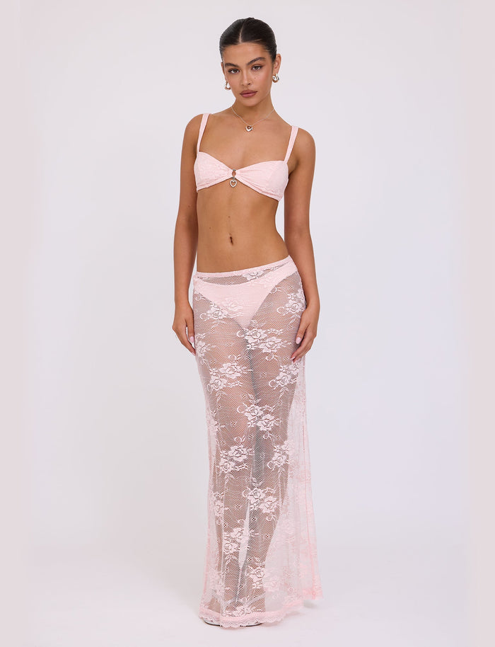 A woman stands in a studio wearing a light pink TIGERMIST bikini top and matching high-waisted bottoms. Over the bottom, she's draped in the XADIE SKIRT - PINK from TIGERMIST, a sheer, floor-length lace skirt with floral patterns. Her hair is pulled back, and she accessorizes with earrings and a necklace.