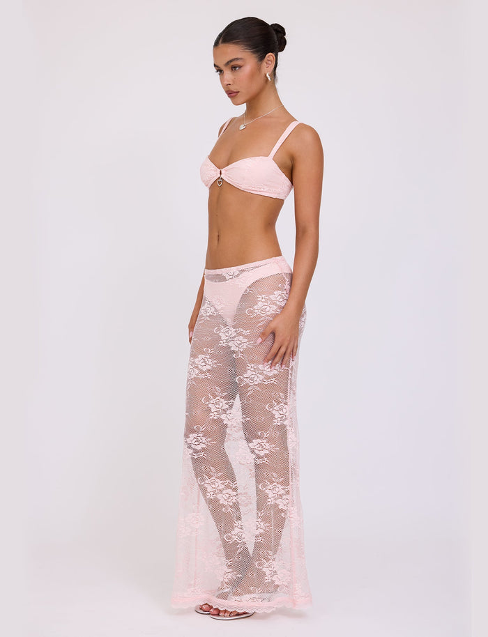 A woman with her hair in a bun is dressed in a matching delicate pink lingerie set from TIGERMIST, featuring a bra, panties, and the XADIE SKIRT in pastel pink. She stands in a profile pose against a plain white background.