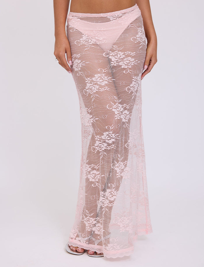 A person in a sheer, floor-length Xadie Skirt by TIGERMIST in pastel pink with floral patterns over matching pale pink underwear. The delicate, flowing outfit showcases intricate lace details against a plain white background.