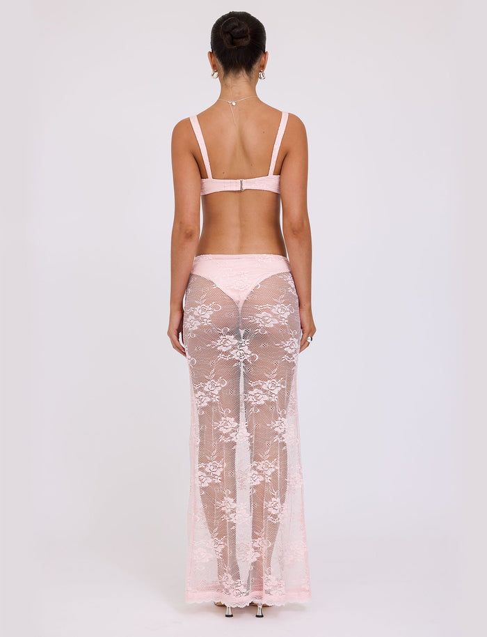 A woman stands with her back to the camera, showcasing a delicate lace bralette from TIGERMIST paired with matching high-waisted panties and the Xadie Skirt in pastel pink. Her hair is styled in a neat bun, highlighting the intricate details of the TIGERMIST lace garments and emphasizing the back view of her outfit.