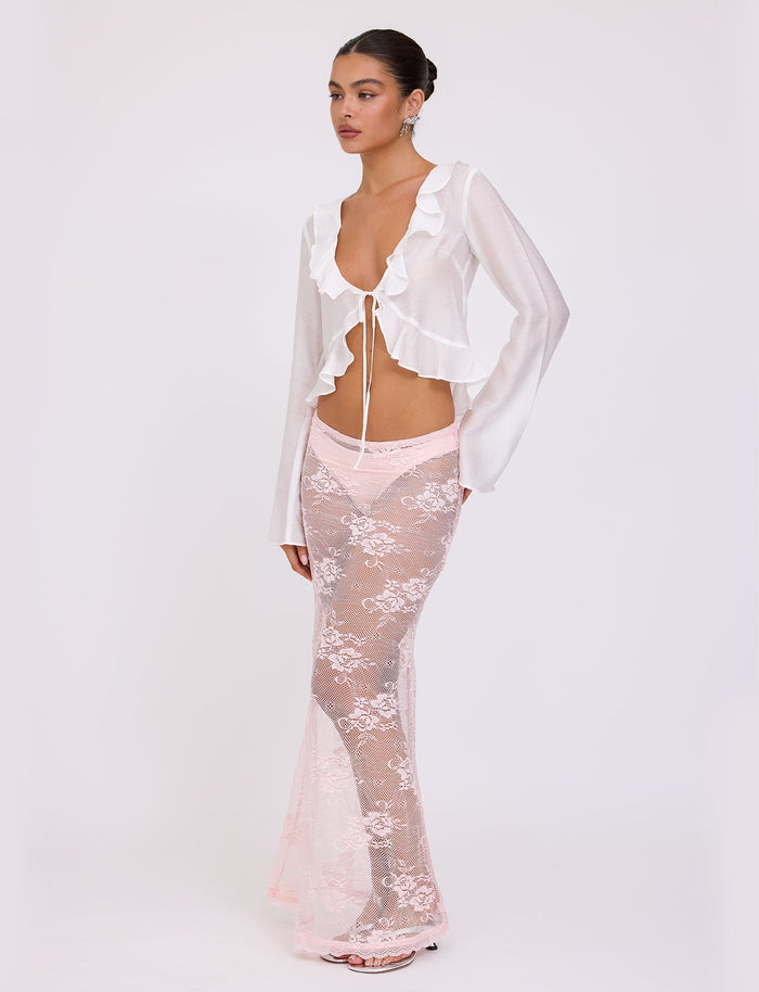 A woman is standing against a plain background, elegantly dressed in a white, long-sleeved ruffled top with a front tie and the XADIE SKIRT from TIGERMIST—a sheer, floor-length pastel pink skirt featuring white floral lace patterns over pale pink undergarments. Her hair is pulled back, and she completes the look with white heels.