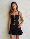A woman with long brown hair stands against a light-colored wall, wearing a stylish black Xandra Skirt by Tiger Mist, featuring a lace-up front and grommet details. The skirt is fitted at the waist and has a ruffled hem. She accessorizes with pearl earrings and has a neutral expression.