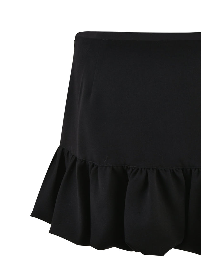 The XANDRA SKIRT - BLACK from TIGERMIST is a black miniskirt with a wide, ruffled hem. The skirt features a fitted upper section that flares out at the ruffle and is made from fabric that appears smooth and slightly shiny.