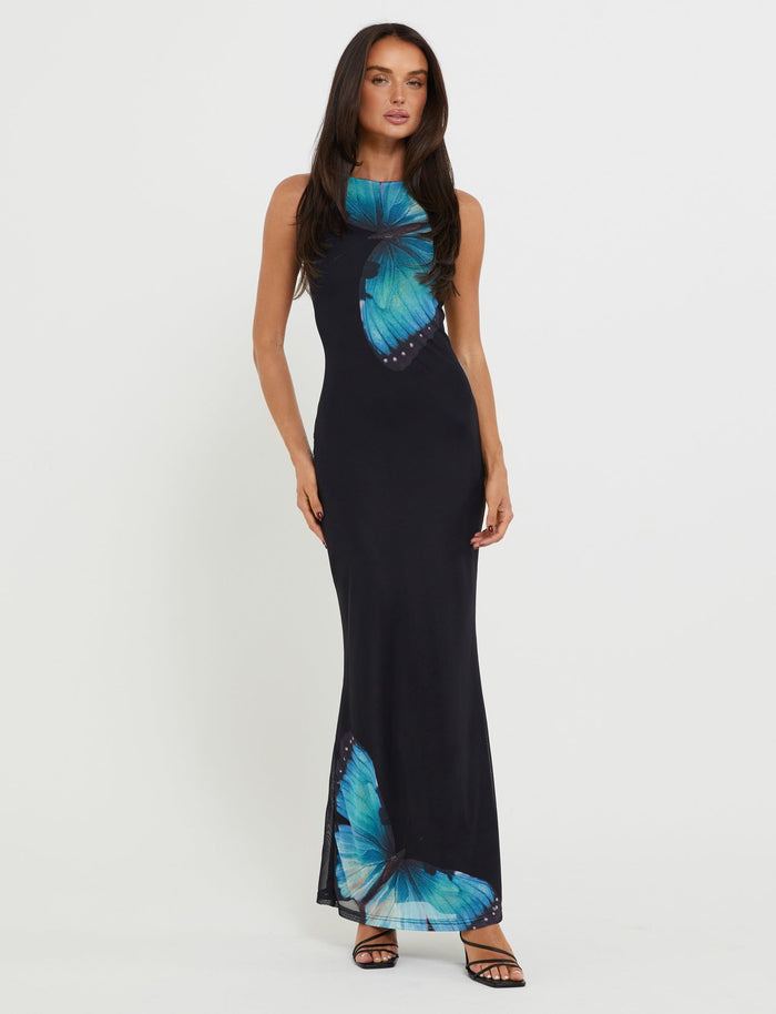A woman stands against a plain white background, wearing the TIGERMIST ZAHRAA DRESS - BLACK : BUTTERFLY, a sleeveless, floor-length black dress adorned with large blue butterfly prints. She has long, straight hair and is wearing black high-heeled sandals, with one leg slightly bent.