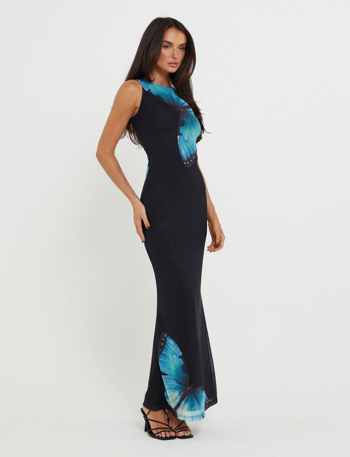 A woman with long dark hair is standing against a plain white background, wearing the TIGERMIST ZAHRAA DRESS in black, which features large blue butterfly prints. She is accessorized with black strappy high heels and is posing with one hand on her hip.