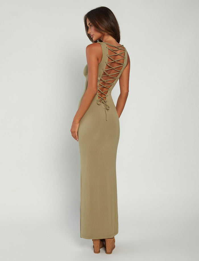 A woman with long brown hair stands facing sideways and looks backward. Clad in a stylish TIGERMIST ZAHRAA Dress in moss green, featuring crisscross lacing down the back, she pairs it effortlessly with high-heeled, open-toed shoes. The background is plain white.
