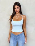 A woman with long brown hair is wearing the ZAIDA TOP in pastel blue from TIGERMIST, along with matching light blue jeans. She has a heart-shaped necklace and is standing against a light beige wall, looking directly at the camera.