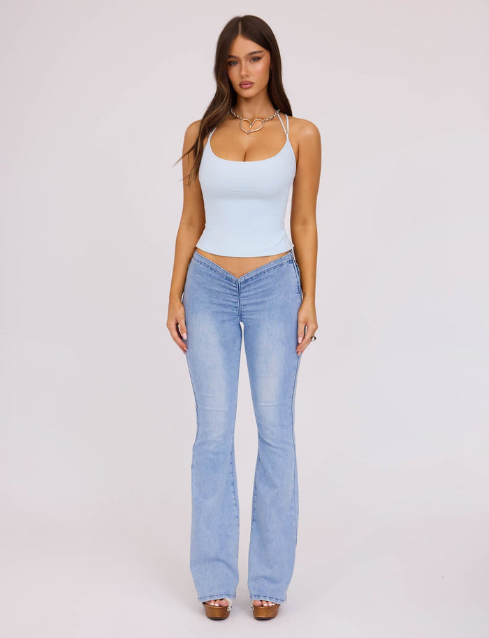 A woman with long hair stands against a plain white background, wearing the ZAIDA TOP - PASTEL BLUE and matching light blue flared jeans from TIGERMIST. She accessorizes with a necklace and open-toe shoes. Her hands are relaxed by her sides, and she looks directly at the camera.