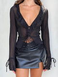 A woman is shown from the shoulders to the thighs wearing the ZELDA TOP - BLACK by TIGERMIST, a sheer black blouse with lace detailing and a deep V-neckline. She pairs it with a black leather mini skirt that has a small front slit, creating an elegant ensemble. The chic Tiger Mist outfit features long sleeves with tie details at the cuffs.