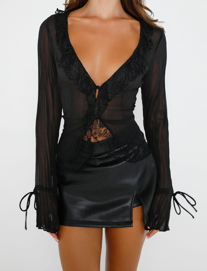 A woman is wearing the TIGERMIST ZELDA TOP - BLACK, a sheer long-sleeve top with lace details, plunging neckline, and tied cuffs. She pairs it with a high-waisted black leather mini skirt from Tiger Mist featuring a small slit on the side. The background is plain white.