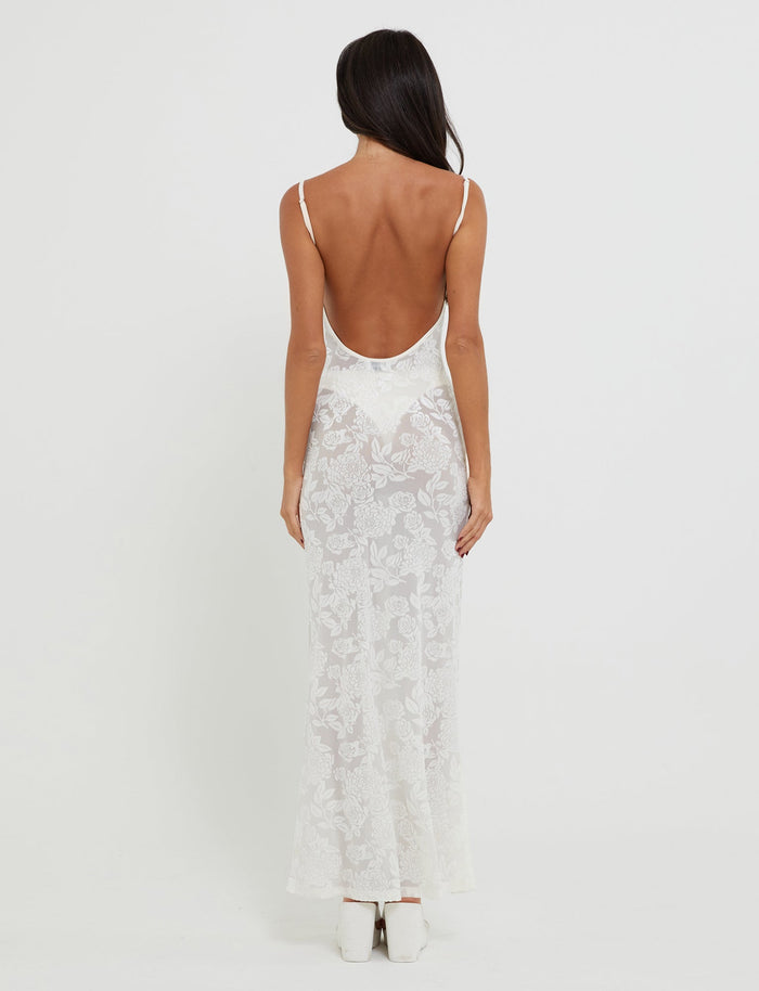 A woman with long dark hair stands with her back to the camera, wearing the ZUZANNA DRESS - WHITE BURNOUT from TIGERMIST. The dress features a low-cut back and thin straps, semi-sheer white lace material. She is also wearing white shoes. The background is plain and white.